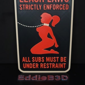 May include: A black and white sign with red text that reads "LEASH LAWS STRICTLY ENFORCED". The sign features a red silhouette of a woman with a leash around her neck. The text below the image reads "ALL SUBS MUST BE UNDER RESTRAINT".