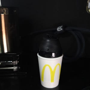 May include: A black electronic device sits atop a white paper cup with a yellow "M" logo. The device has a rounded top with a textured design. The cup is on a dark surface.