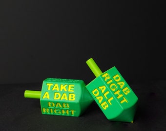 Könnte beinhalten: Zwei grüne Dreidels mit gelber Beschriftung. Ein Dreidel lautet "Take a Dab Dab Right" und der andere lautet "Dab Right All Dab".