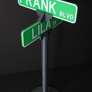 May include: A green street sign with the text "FRANK BLVD" and a smaller green street sign with the text "LILA ST" attached to it. The signs are mounted on a black metal stand.