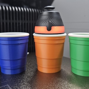 May include: An orange, black, and white portable beverage dispenser with three colorful cups. The cups are blue, orange, and green, each with a white rim. The dispenser has a black top and an orange light.