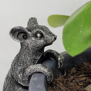 May include: A silver-coloured mouse figurine perched on the edge of a black planter pot. The mouse has a detailed, realistic design with a textured surface.