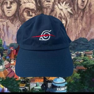 Può includere: Un cappello da baseball blu scuro con un logo ricamato bianco e rosso. Il logo è un vortice stilizzato con una linea rossa che lo attraversa.