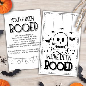 Könnte beinhalten: Zwei Halloween-Karten mit den Sätzen "You've Been Booed" und "We've Been Booed" in Schwarz. Die Karten zeigen Geister- und Fledermaus-Illustrationen. Kürbisse, Spinnen und eine Skeletthand sind um die Karten herum verstreut.