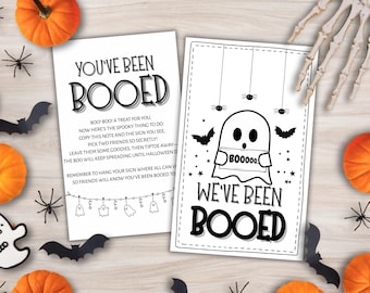 May include: Two Halloween-themed cards with the phrases "You've Been Booed" and "We've Been Booed" in black lettering. The cards feature ghost and bat illustrations. Pumpkins, spiders, and a skeleton hand are scattered around the cards.