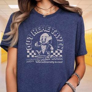 Puede incluir: Una camiseta azul con un gr&aacute;fico de un cono de helado de dibujos animados con una cara y el texto "Out Here Tryin' Not To Melt Down" y "Neurodiversity is cool".