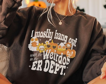 May include: Brown sweatshirt with a graphic print of a ghost, pumpkins, and flowers. The text on the sweatshirt reads "I mostly hang out with the weirdos in the ER DEPT."