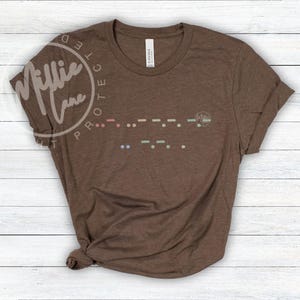 May include: A brown t-shirt with rolled-up sleeves. The shirt features a Morse code design in various colors across the chest. The brand name "Millie Lane" is printed on the upper left side of the shirt.