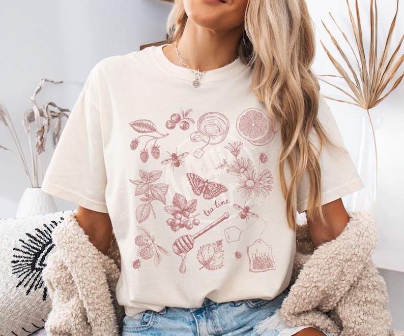 May include: Cream-colored t-shirt featuring a pink illustration of tea-related elements. The design includes a teacup, tea bag, honey dipper, berries, flowers, a butterfly, and the words "tea time". The shirt is a comfortable, casual style.