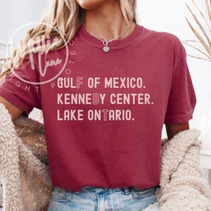 May include: A maroon short-sleeve crewneck t-shirt featuring white text printed on the front that reads GULF OF MEXICO. KENNEDY CENTER. LAKE ONTARIO. The shirt has a relaxed fit and is paired with blue denim jeans and a textured beige cardigan.