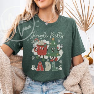 May include: A dark green short-sleeve t-shirt featuring a graphic of a red mug with a candy cane and a green Christmas tree with faces. The text reads Jingle Bells & ADLs in a retro font, surrounded by white snowflakes on a white background.