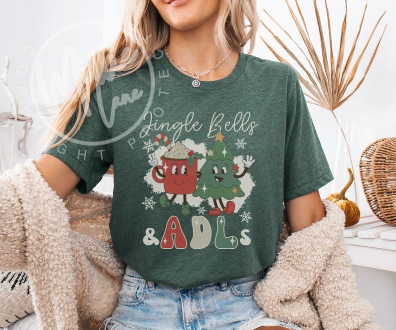 May include: A dark green short-sleeve t-shirt featuring a graphic of a red mug with a candy cane and a green Christmas tree with faces. The text reads Jingle Bells & ADLs in a retro font, surrounded by white snowflakes on a white background.