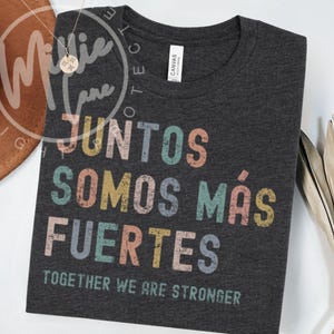 Könnte beinhalten: Ein dunkelgraues T-Shirt mit Rundhalsausschnitt und dem Text JUNTOS SOMOS MÁS FUERTES sowie TOGETHER WE ARE STRONGER in einer verwaschenen, mehrfarbigen Schriftart. Das Oberteil verfügt über ein weißes Markenetikett am inneren Halsausschnitt.