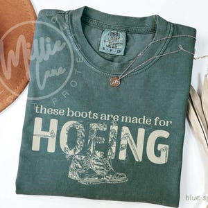 Puede incluir: Una camiseta azul abeto con el texto "these boots are made for HOEING" en crema. La palabra "HOEING" presenta un diseño floral. Un collar plateado con un colgante está sobre la camiseta.
