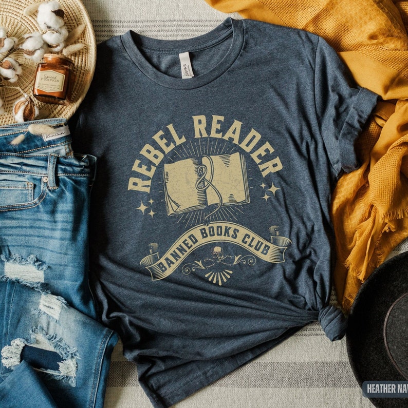 K&ouml;nnte beinhalten: Ein dunkelblaues T-Shirt mit einem Vintage-Grafikdesign, auf dem "Rebel Reader Banned Books Club" steht. Die Grafik zeigt ein offenes Buch mit einem Lesezeichenband und einem Totenkopf mit Knochen.