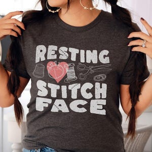 May include: A dark grey t-shirt with the text "Resting Stitch Face" in white with a heart and sewing-themed imagery.