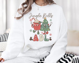 May include: A white crewneck sweatshirt featuring a graphic print of a red mug with a candy cane and a green Christmas tree with faces. The text Jingle Bells & ADLs is printed in brown, red, and green. The design includes small red and green snowflakes.