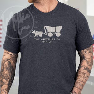 Puede incluir: Camiseta gris oscuro con una imagen pixelada de una carreta cubierta tirada por un buey, con el texto blanco "YOU LISTENED TO RFK JR". La camiseta es de manga corta.