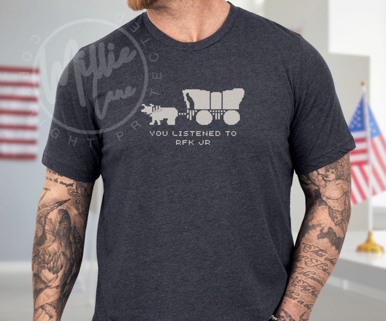 Puede incluir: Camiseta gris oscuro con una imagen pixelada de una carreta cubierta tirada por un buey, con el texto blanco "YOU LISTENED TO RFK JR". La camiseta es de manga corta.