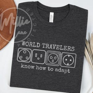 May include: Dark gray t-shirt with the text "WORLD TRAVELERS know how to adapt" and illustrations of international electrical outlet plugs in white. The shirt is made of a soft material.