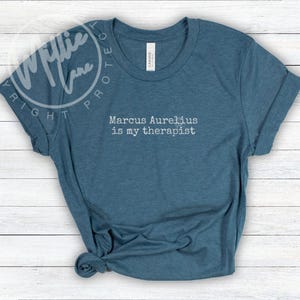 以下が含まれることがあります： 「Marcus Aurelius is my therapist」という白文字がプリントされた青いTシャツ。袖は折り返され、裾は結ばれています。柔らかい素材で作られています。