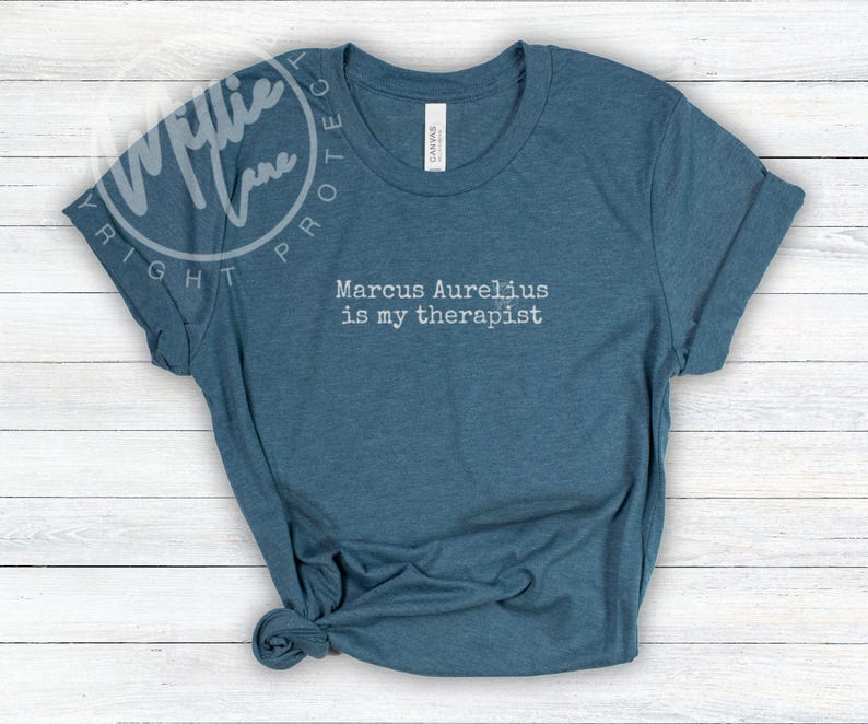 以下が含まれることがあります： 「Marcus Aurelius is my therapist」という白文字がプリントされた青いTシャツ。袖は折り返され、裾は結ばれています。柔らかい素材で作られています。