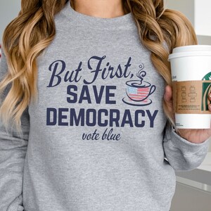 May include: A gray sweatshirt with the text "But First, Save Democracy vote blue" in navy blue. The text is accompanied by a stylized coffee cup with an American flag design.