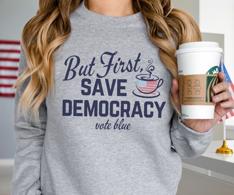 May include: A gray sweatshirt with the text "But First, Save Democracy vote blue" in navy blue. The text is accompanied by a stylized coffee cup with an American flag design.