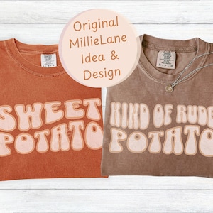 May include: Two t-shirts with retro text designs. One is burnt orange with "SWEET POTATO" in light orange. The other is brown with "KIND OF RUDE POTATO" in light orange. A silver necklace with a pendant is on the brown shirt.