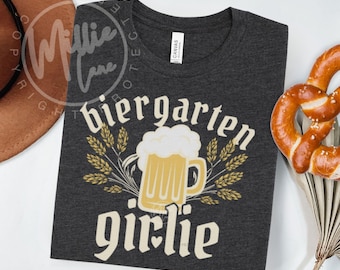 Op de afbeelding: Donkergrijs T-shirt met de woorden "Biergarten Girlie" in een vintage lettertype, met een bierpul-afbeelding. Het shirt is opgevouwen, met een pretzel en een hoed op de achtergrond. Het shirt is gemaakt van een zacht materiaal.