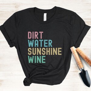 May include: Black t-shirt featuring the words "DIRT WATER SUNSHINE WINE" in pink, blue, beige, and green block letters. Gardening tools, a trowel and a hand rake, are placed near the shirt. The shirt is knotted at the bottom.