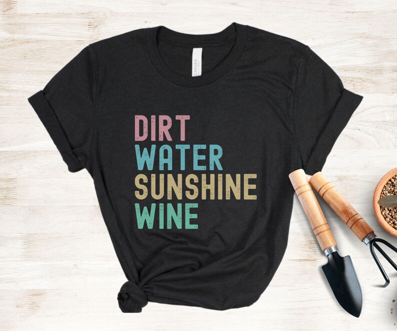 May include: Black t-shirt featuring the words "DIRT WATER SUNSHINE WINE" in pink, blue, beige, and green block letters. Gardening tools, a trowel and a hand rake, are placed near the shirt. The shirt is knotted at the bottom.