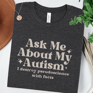 May include: Dark grey t-shirt with the text "Ask Me About My Autism" in a beige, stylized font. Below the text is the phrase "I destroy pseudoscience with facts". The shirt is made of a soft material.