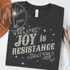 May include: Dark gray t-shirt with the words "JOY is RESISTANCE" in a vintage-style font. The design includes floral and star accents. The shirt is made of a soft material.