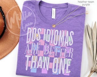 May include: A heather team purple t-shirt featuring a white graphic print that reads "DOS IDIOMAS ARE BETTER THAN ONE" over a checkered pattern of light pink, light blue, and light yellow squares. A gold-toned necklace rests on the neckline.