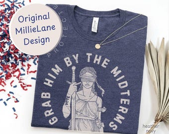 以下が含まれることがあります： ヘザーネイビーのTシャツ。剣を持つ正義の女神のグラフィックが描かれ、手首には「VOTE」の文字がある。図形の周囲には「GRAB HIM BY THE MIDTERMS」という文字が円状にプリントされている。襟元には細いシルバーのネックレスが置かれている。