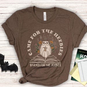 May include: Brown t-shirt with a whimsical ghost design. The ghost, wearing glasses and a witch hat, is reading a book and holding a drink. Text reads "Came for the Heebies, Stayed for the Jeebies". A bat and a book are in the background.