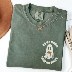 Peut inclure: T-shirt vert olive &agrave; col rond et manches courtes. Le t-shirt pr&eacute;sente un motif de fant&ocirc;me tenant une tasse de caf&eacute;, avec le texte "DO NO HARM TAKE NO SHEET". Un collier en or avec un pendentif est sur le t-shirt.
