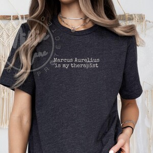 以下が含まれることがあります： 「Marcus Aurelius is my therapist」の白文字がプリントされた濃いグレーのTシャツ。半袖でクルーネック。生地は柔らかく快適そうです。