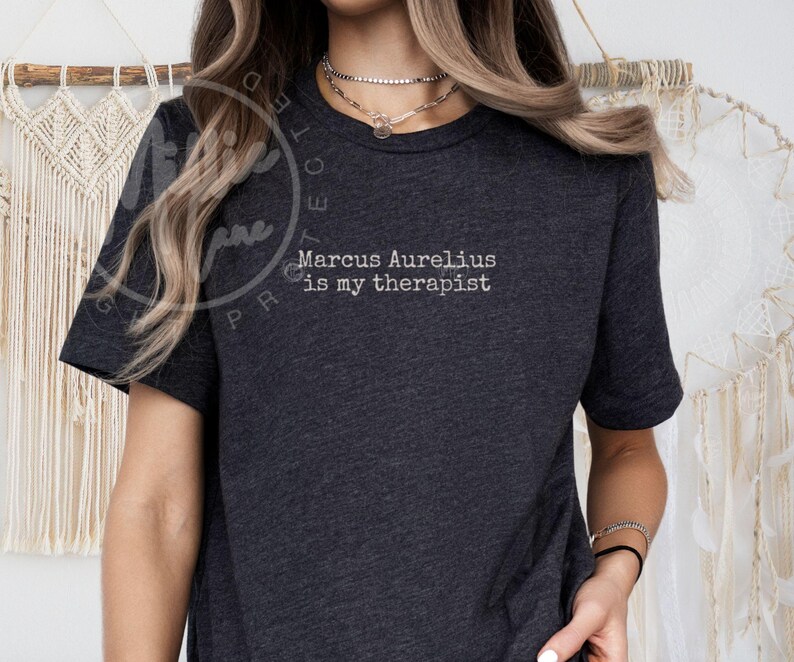 以下が含まれることがあります： 「Marcus Aurelius is my therapist」の白文字がプリントされた濃いグレーのTシャツ。半袖でクルーネック。生地は柔らかく快適そうです。