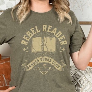 K&ouml;nnte beinhalten: Ein gr&uuml;nes T-Shirt mit dem Text "Rebel Reader Banned Books Club" in einer Vintage-Schriftart. Das Design zeigt ein Buch mit einem Lesezeichen und Sternen.