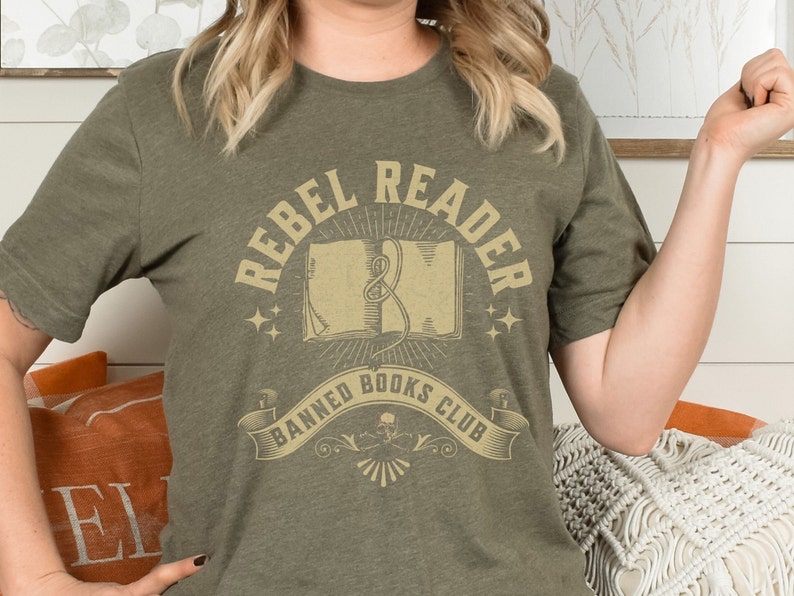 K&ouml;nnte beinhalten: Ein gr&uuml;nes T-Shirt mit dem Text "Rebel Reader Banned Books Club" in einer Vintage-Schriftart. Das Design zeigt ein Buch mit einem Lesezeichen und Sternen.