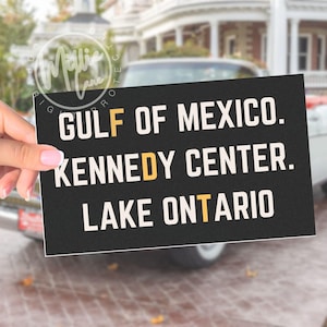 May include: A rectangular black sign with white text reading GULF OF MEXICO, KENNEDY CENTER, and LAKE ONTARIO. The letters F, D, and T are highlighted in a contrasting yellow color, creating a vertical alignment of the letters F, D, and T.