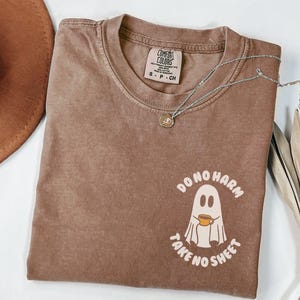 Peut inclure: Un t-shirt marron Comfort Colors &agrave; col rond et manches courtes. Le t-shirt pr&eacute;sente un motif de fant&ocirc;me tenant une tasse de caf&eacute;, avec le texte "DO NO HARM TAKE NO SHEET". Un collier argent&eacute; avec un pendentif est pos&eacute; sur le t-shirt.