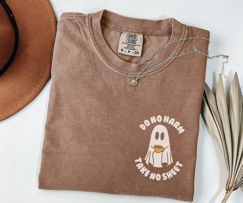 Peut inclure: Un t-shirt marron Comfort Colors &agrave; col rond et manches courtes. Le t-shirt pr&eacute;sente un motif de fant&ocirc;me tenant une tasse de caf&eacute;, avec le texte "DO NO HARM TAKE NO SHEET". Un collier argent&eacute; avec un pendentif est pos&eacute; sur le t-shirt.