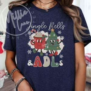 May include: A navy blue short-sleeve t-shirt featuring a graphic print of a red mug with a candy cane and a green Christmas tree with the text Jingle Bells & ADLs. The design includes white snowflakes and cartoon-style faces on the mug and tree.