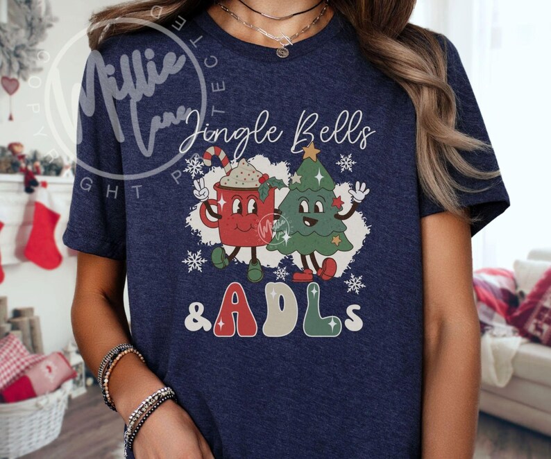 May include: A navy blue short-sleeve t-shirt featuring a graphic print of a red mug with a candy cane and a green Christmas tree with the text Jingle Bells & ADLs. The design includes white snowflakes and cartoon-style faces on the mug and tree.