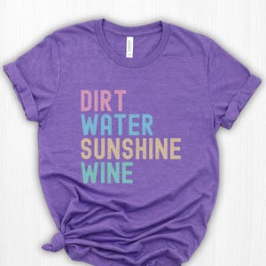 May include: A purple t-shirt with the words "DIRT WATER SUNSHINE WINE" stacked in different colours: pink, blue, beige, and teal. The shirt has a crew neck and short sleeves.