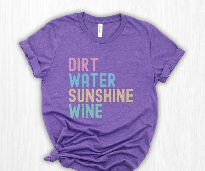 May include: A purple t-shirt with the words "DIRT WATER SUNSHINE WINE" stacked in different colours: pink, blue, beige, and teal. The shirt has a crew neck and short sleeves.