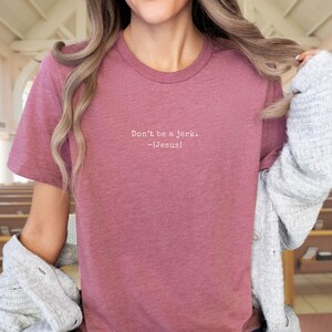 Peut inclure: Un t-shirt rose poudr&eacute; avec l'inscription blanche "Don't be a jerk. -[Jesus]" sur le devant. Le t-shirt est de style col rond et semble &ecirc;tre fait d'une mati&egrave;re douce. L'arri&egrave;re-plan montre un int&eacute;rieur d'&eacute;glise.